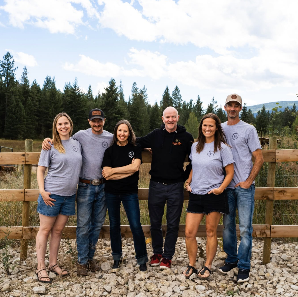 Group of six people posing outdoors with a wooden fence and forest in the background- yankee flat meats british columbia